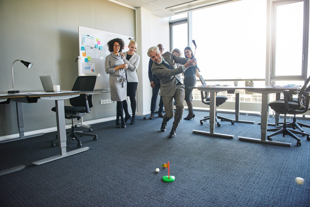Diverse group of laughing coworkers watching a mature colleague swinging a toy golf club during a team building exercise in a modern office